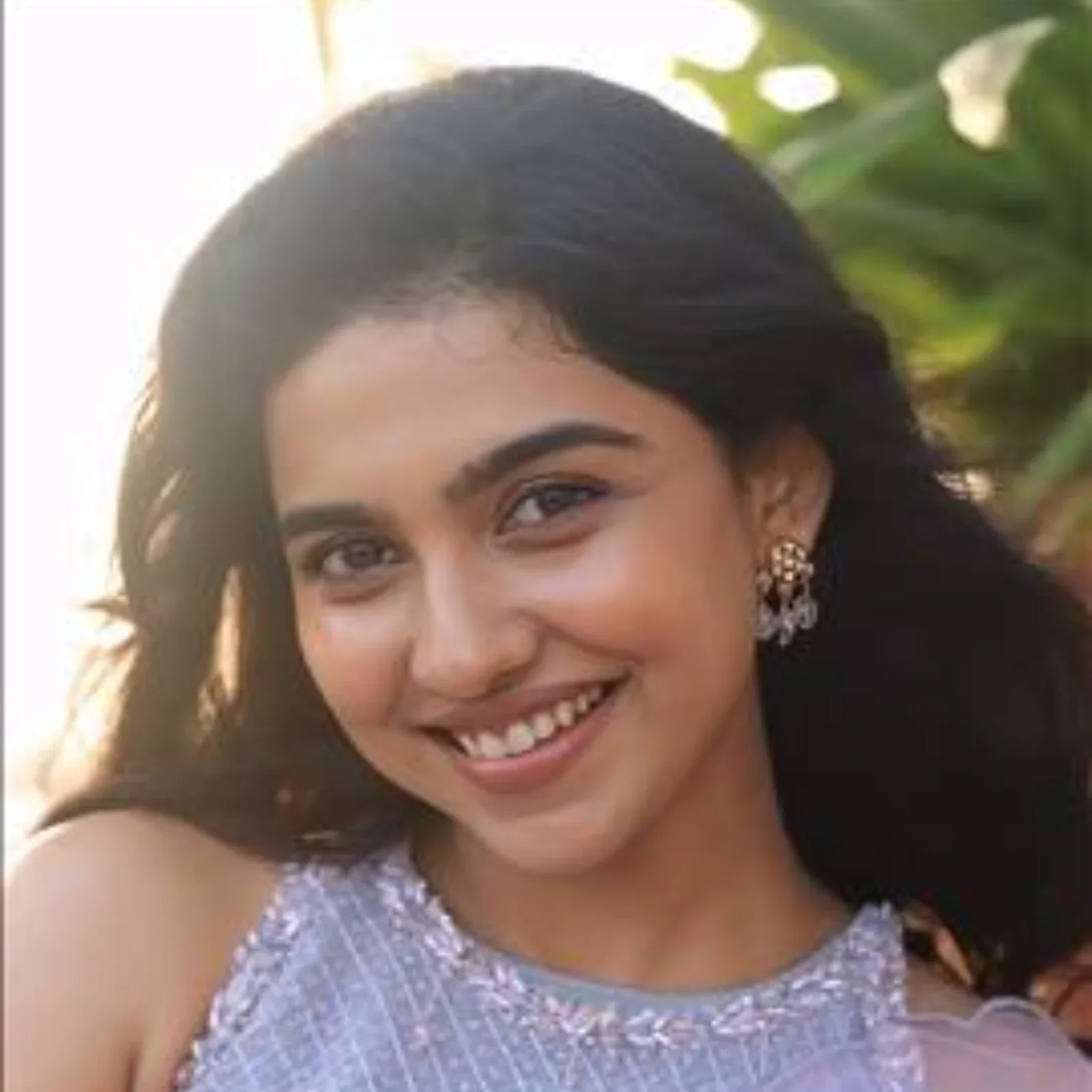 Close-up portrait of Mamitha Baiju smiling, wearing a lavender embroidered top and chandelier earrings, with a blurred sunny outdoor background.