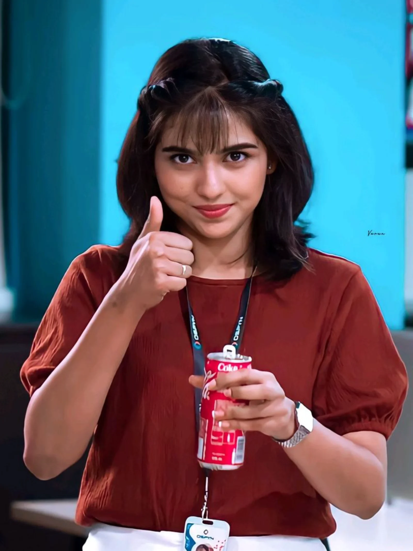 Mamitha Baiju smiling and giving a thumbs-up, holding a red Coca-Cola can, wearing a rust-colored top against a blue background.