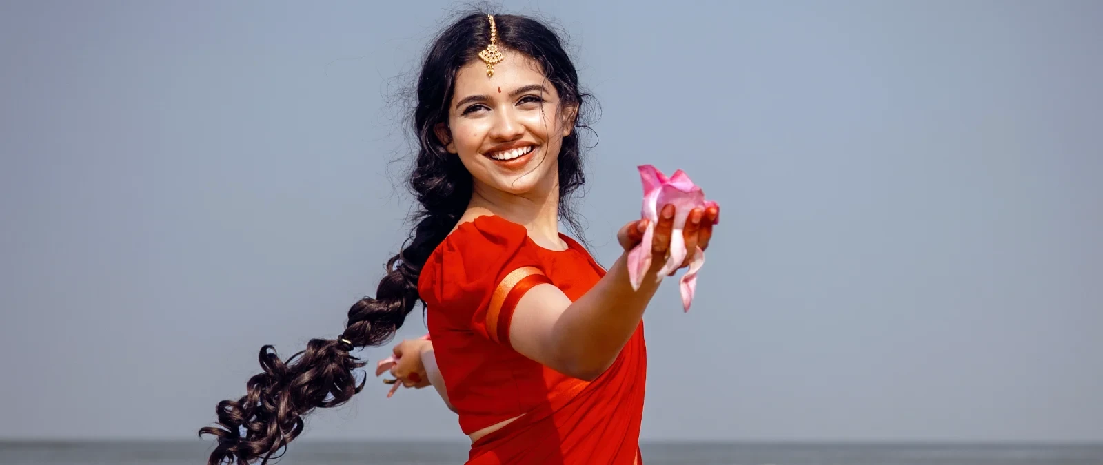 Mamitha Baiju smiling broadly in a red traditional top, holding pink flower petals, with a long curly braid and a maang tikka, against a light blue sky.