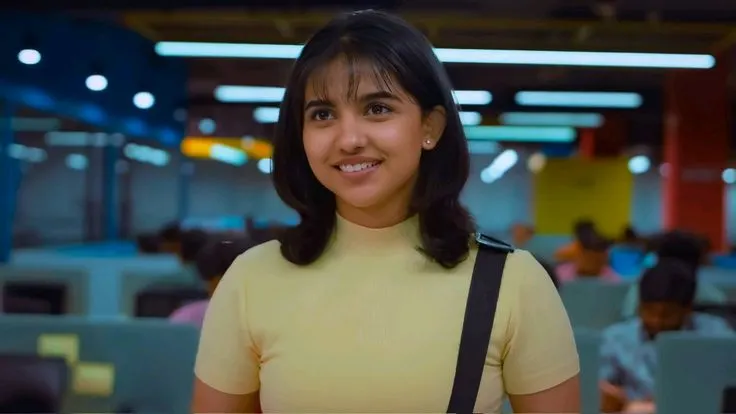 Mamitha Baiju, a young Indian actress, smiling cheerfully in a light yellow high-neck top with dark shoulder-length hair and bangs, in an indoor setting.