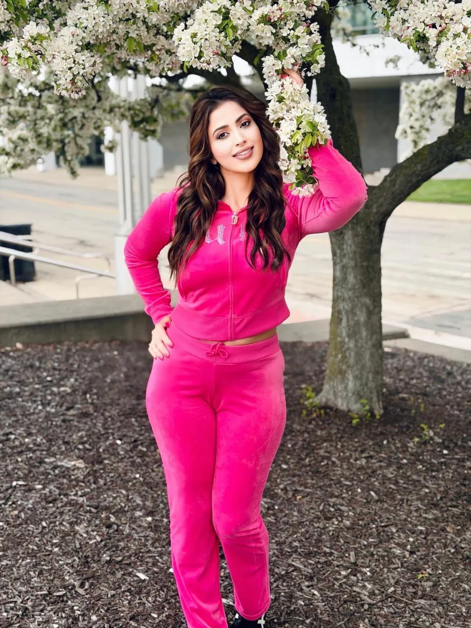 Samaira Sandhu poses charmingly in a bright pink velour tracksuit under a blooming tree. Samaira Sandhu smiling, wearing a bright hot pink velour tracksuit, standing under a tree with white blossoms, with one hand on her hip and the other touching a branch.