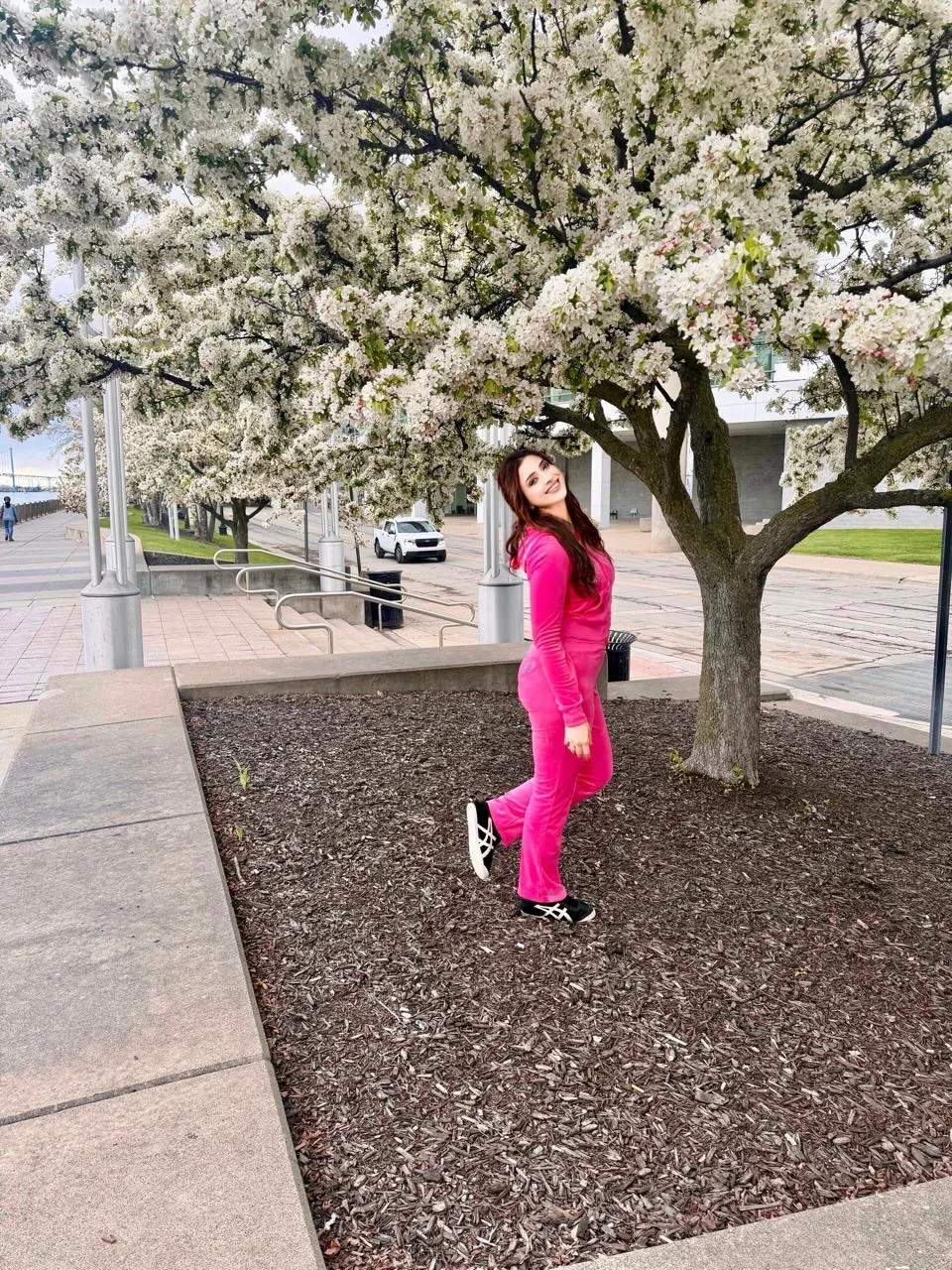 Samaira Sandhu smiling brightly in a hot pink velour tracksuit under a tree with white blossoms.