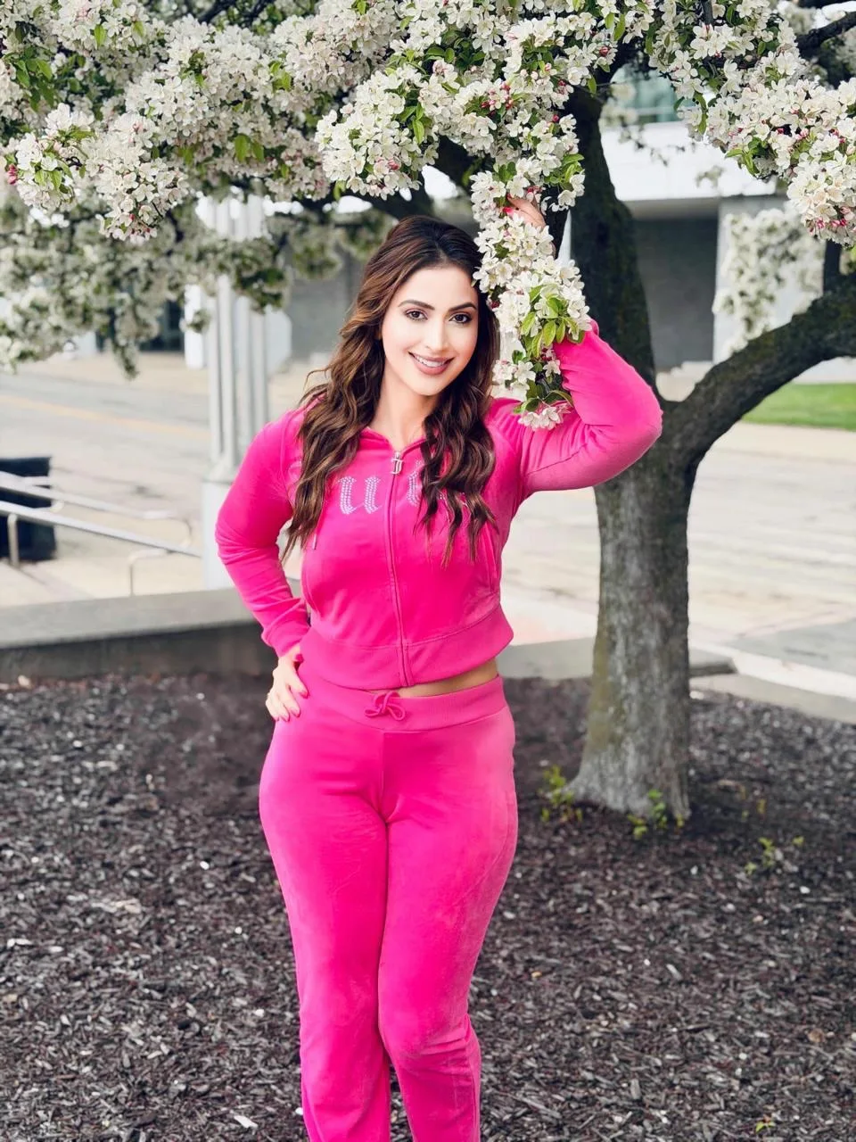 Samaira Sandhu smiling brightly in a hot pink velour tracksuit, standing next to a tree with white blossoms.