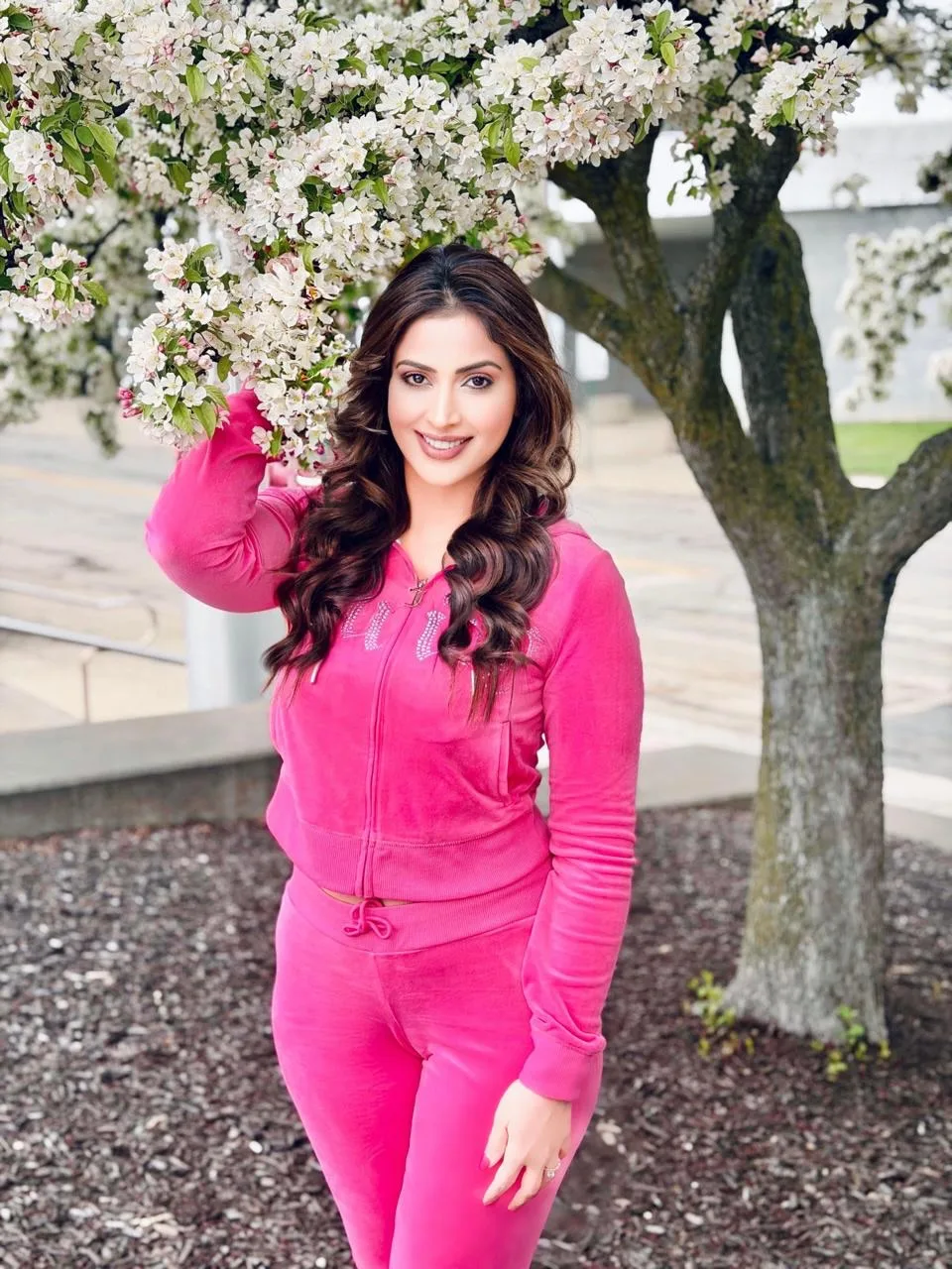 Samaira Sandhu, an Indian actress, smiling brightly in a vibrant pink velour tracksuit, standing next to a tree with white spring blossoms.