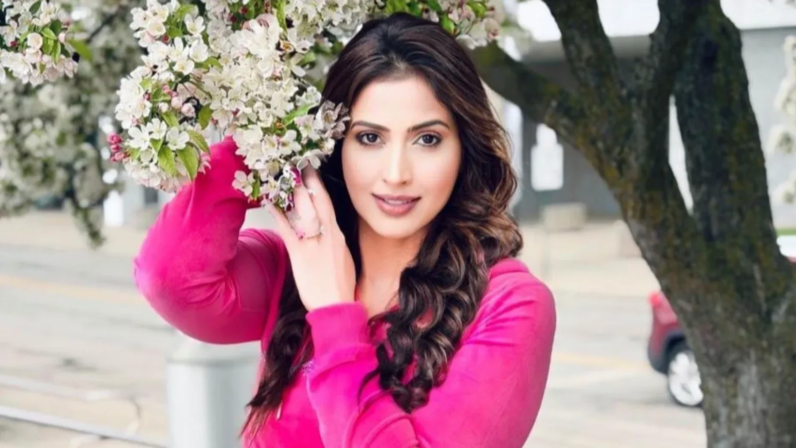 Samaira Sandhu, an Indian actress, smiling while holding white and pink blossoms, wearing a bright pink long-sleeved top.