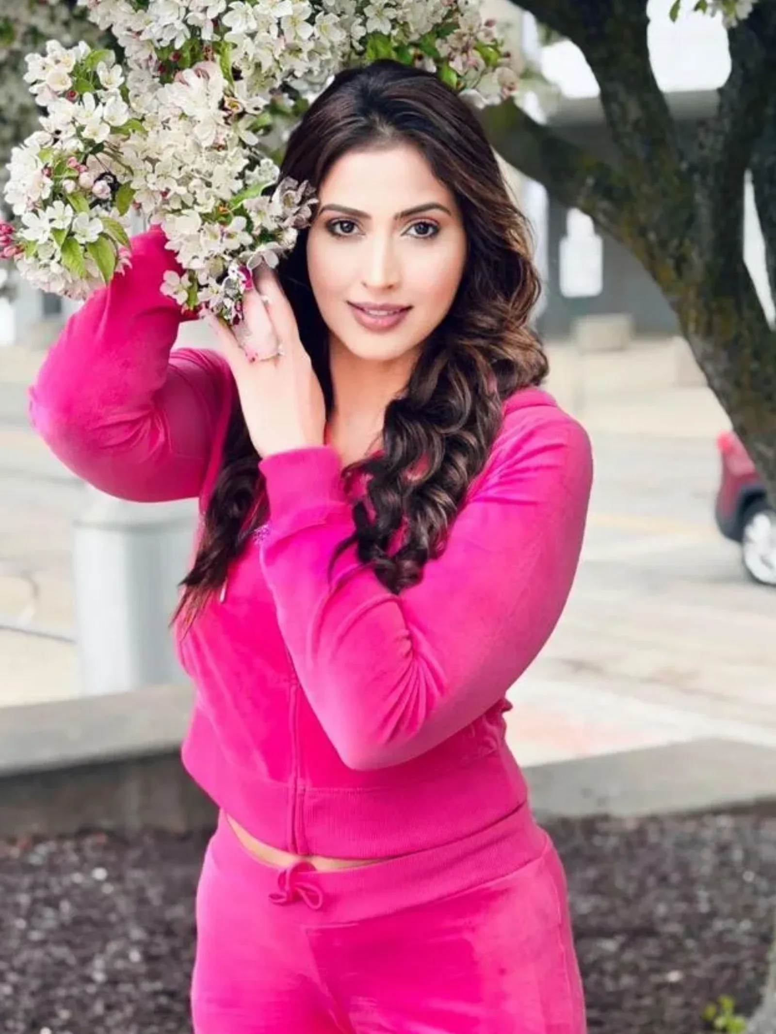 Samaira Sandhu, an Indian actress, smiling while posing outdoors in a bright pink velour tracksuit, holding white cherry blossoms near her face.