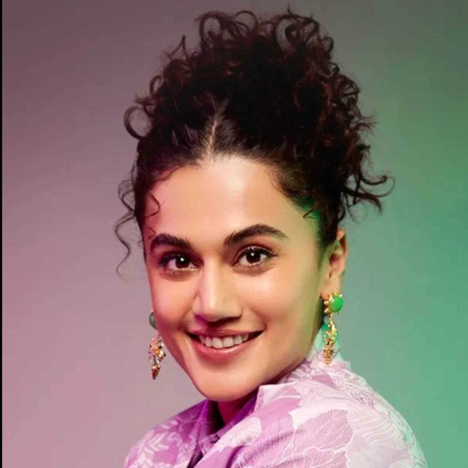 Close-up portrait of Taapsee Pannu smiling, with curly hair tied up, wearing a lavender patterned top and green and gold dangle earrings against a gradient background.