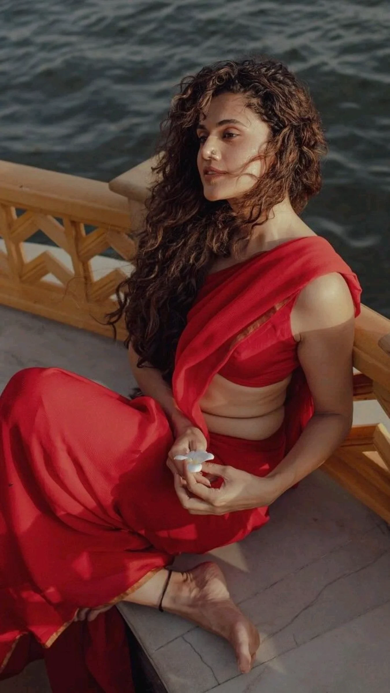 Taapsee Pannu sitting by the water in a red saree, holding a white flower, with her curly hair flowing.