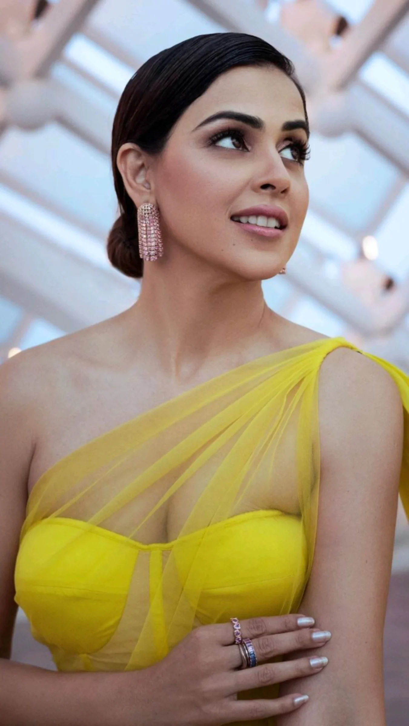 Genelia D'Souza smiling, wearing a bright yellow one-shoulder dress with sheer tulle, pink rhinestone earrings, and her hair in a bun.