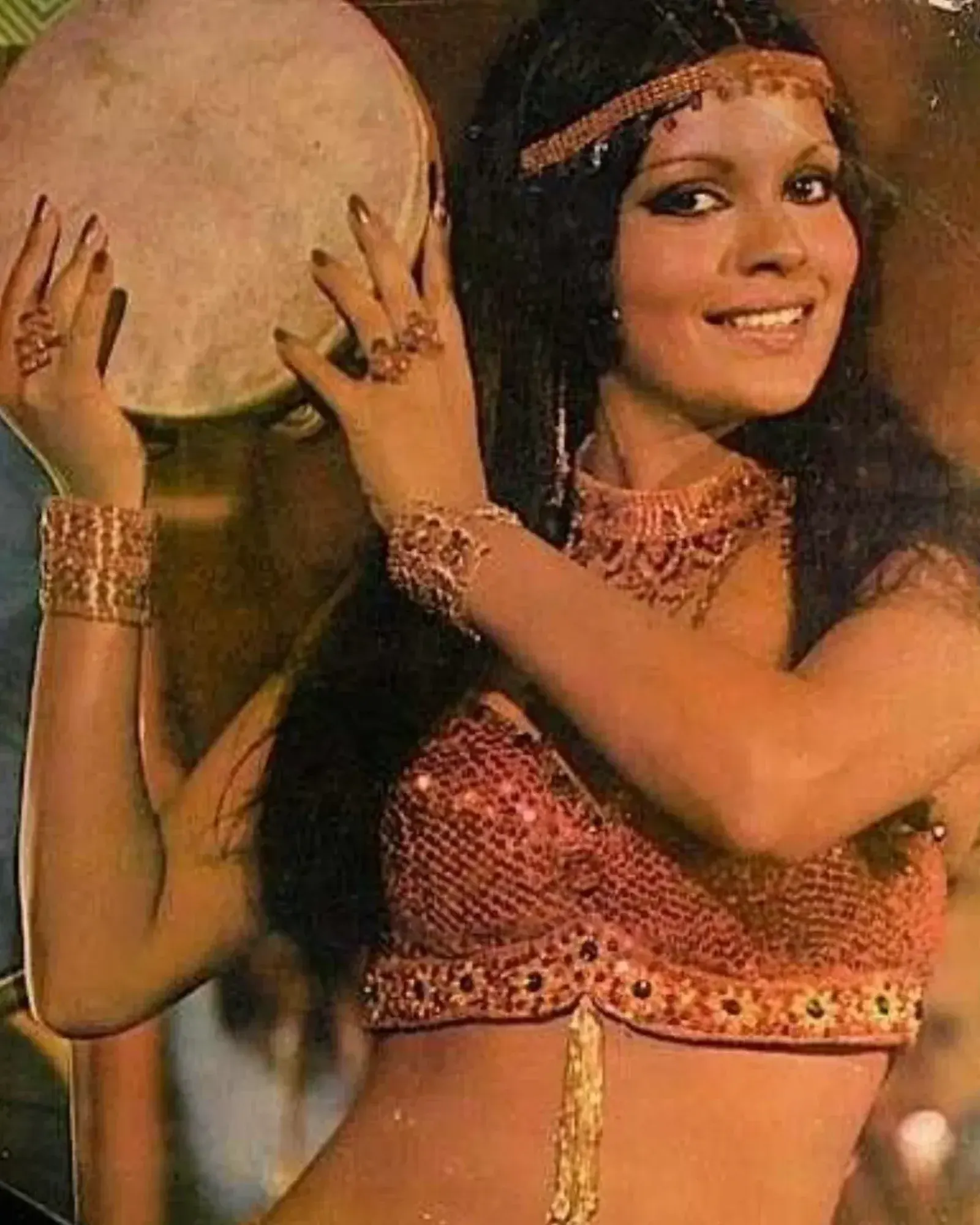 Zeenat Aman performing joyfully with a drum, wearing a sparkling red outfit and jewelry.