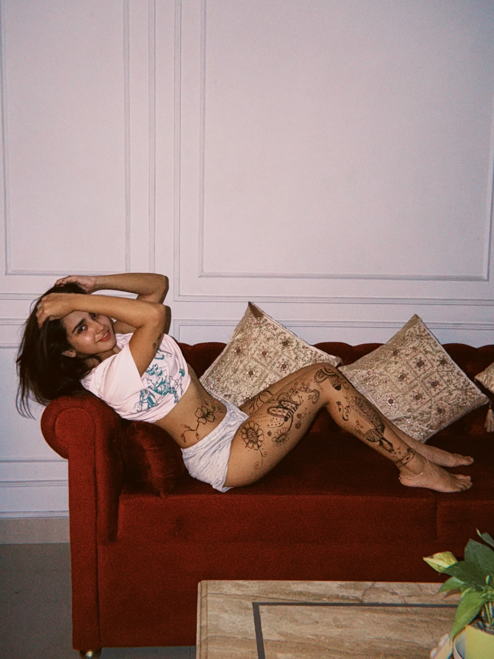 Roshni Walia reclining on a red velvet sofa, smiling at the camera, wearing a pink crop top and shorts, with intricate henna-style body art covering her legs and hip.