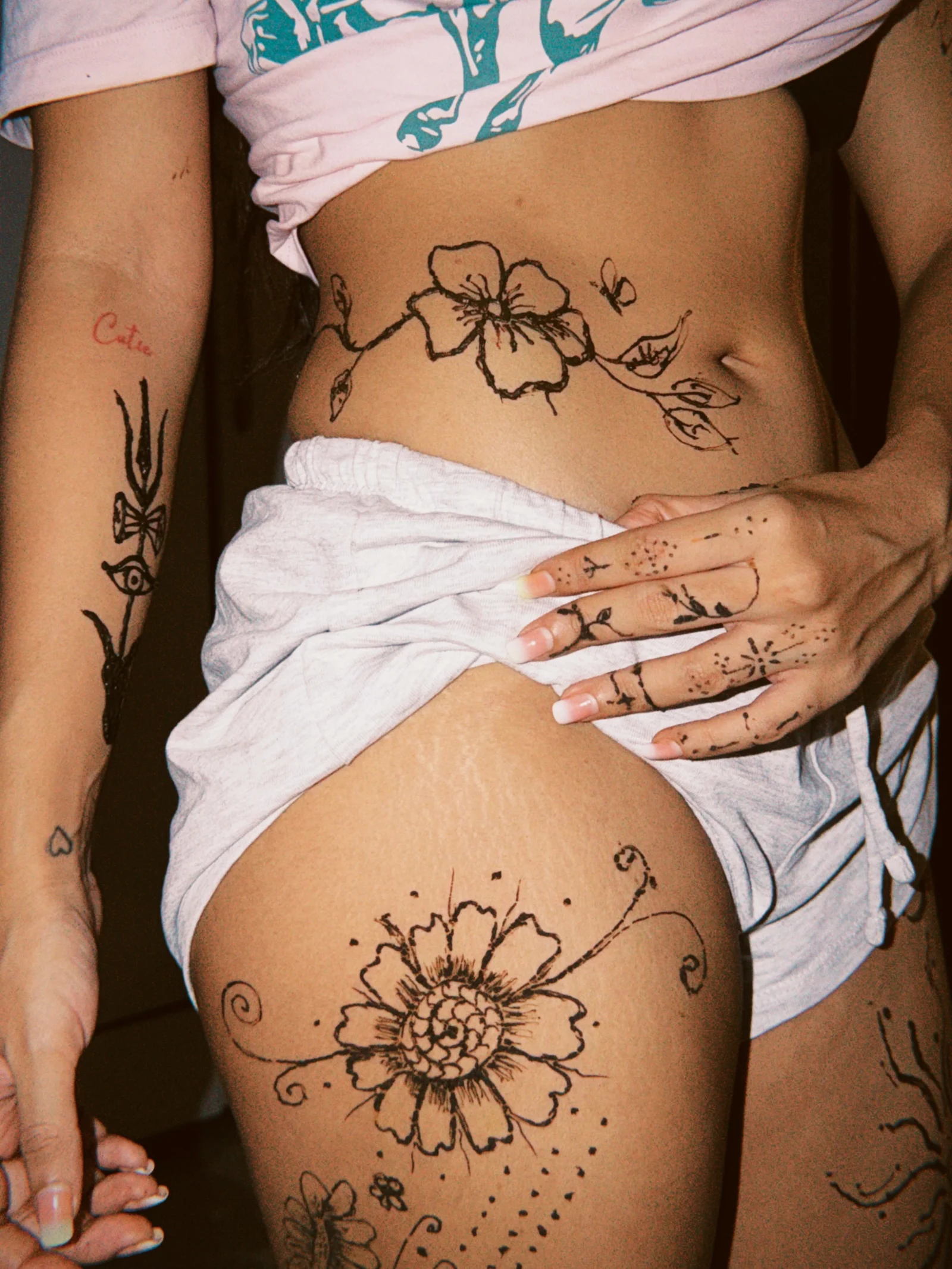 Close-up of Roshni Walia's torso, arm, and thigh, adorned with black henna floral and vine tattoos. Her red 'Cutie' tattoo is visible on her bicep, and she wears a pink top and white shorts.