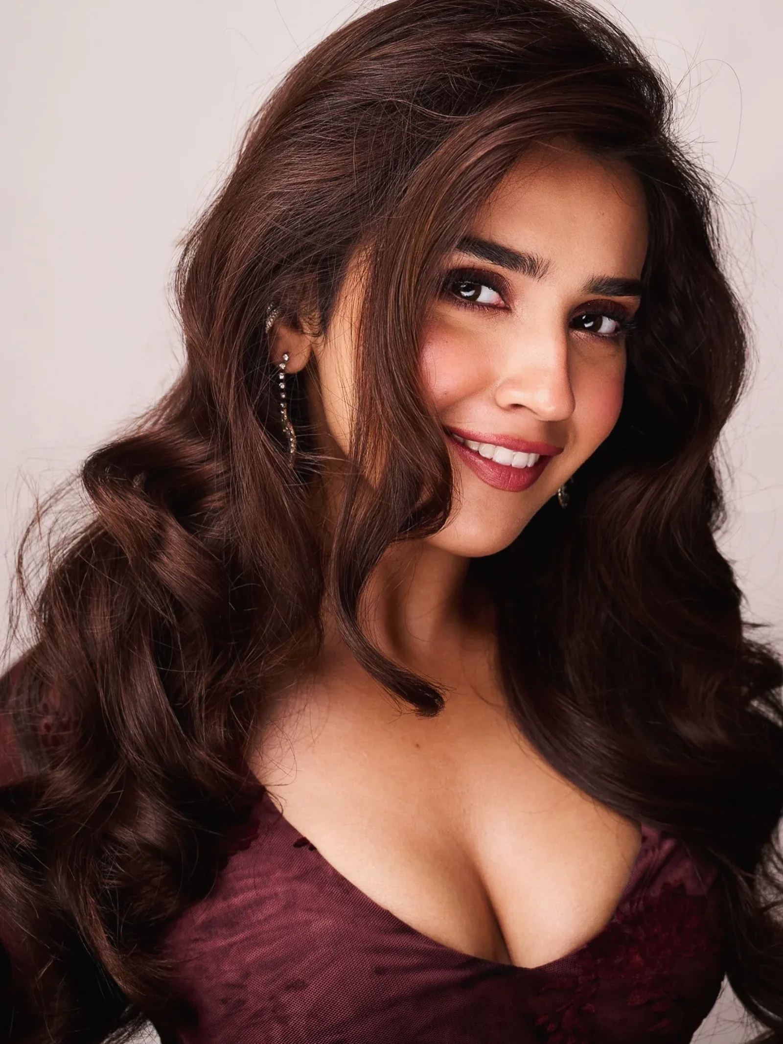 Close-up portrait of Roshni Walia smiling, wearing a maroon top and sparkling drop earrings, with long wavy dark brown hair.