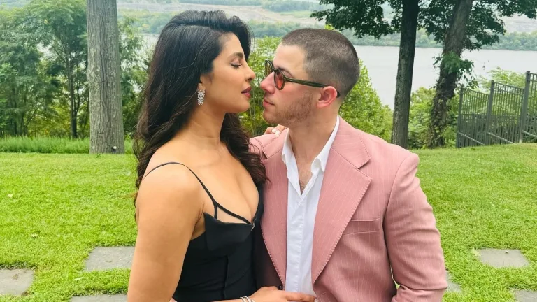 Priyanka Chopra in a black dress and Nick Jonas in a pink suit, looking at each other intimately outdoors with a green, natural background.