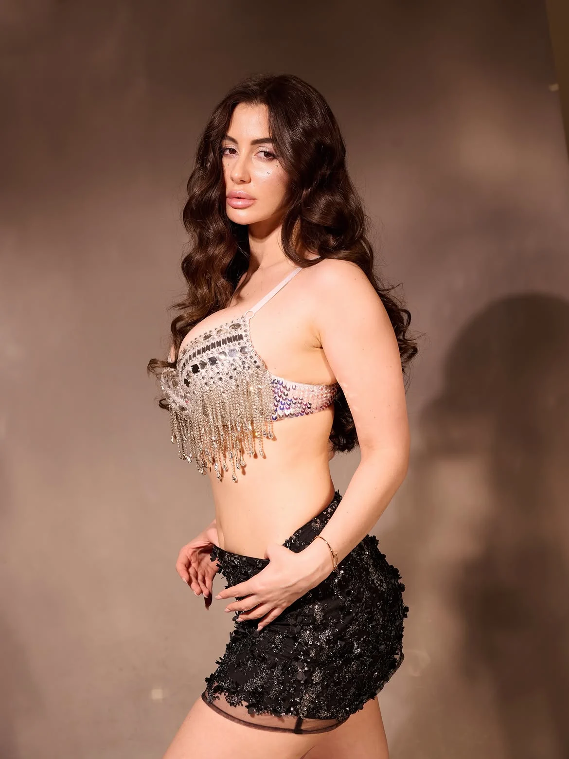 Giorgia Andriani posing in a shimmering silver embellished bra-top with fringe and a black sequined mini-skirt, against a brown background.
