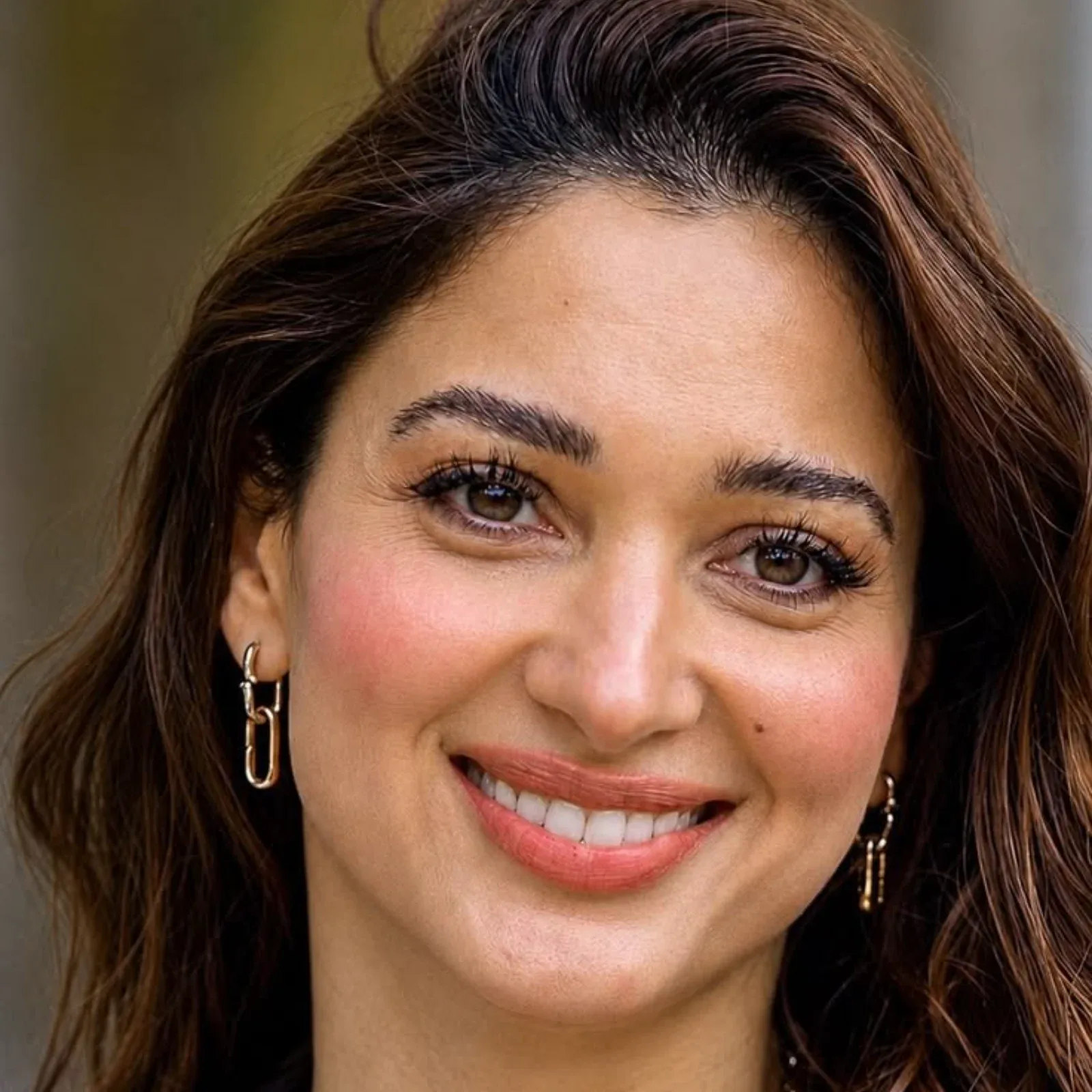 Close-up portrait of Tamannaah Bhatia smiling, showing her brown eyes, long eyelashes, natural makeup, and gold chain-link earrings.