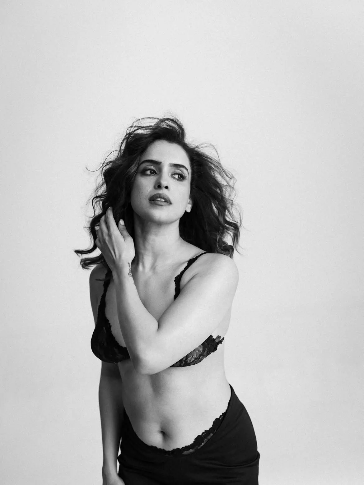 Black and white portrait of Sanya Malhotra wearing a black lace bralette and high-waisted bottoms, with windswept dark hair, looking upwards with a pensive expression.
