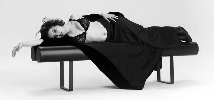 Sanya Malhotra lying on a black bench, wearing a black lace bralette and flowing black fabric, looking sultrily at the camera in a black and white photo.