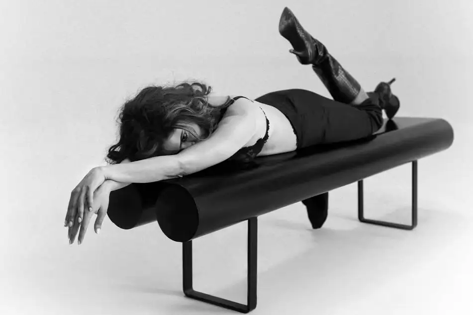 Black and white image of Sanya Malhotra lying on a black cylindrical bench, wearing a bra and high-heeled boots, with her wavy hair spread around her head, looking towards the camera.