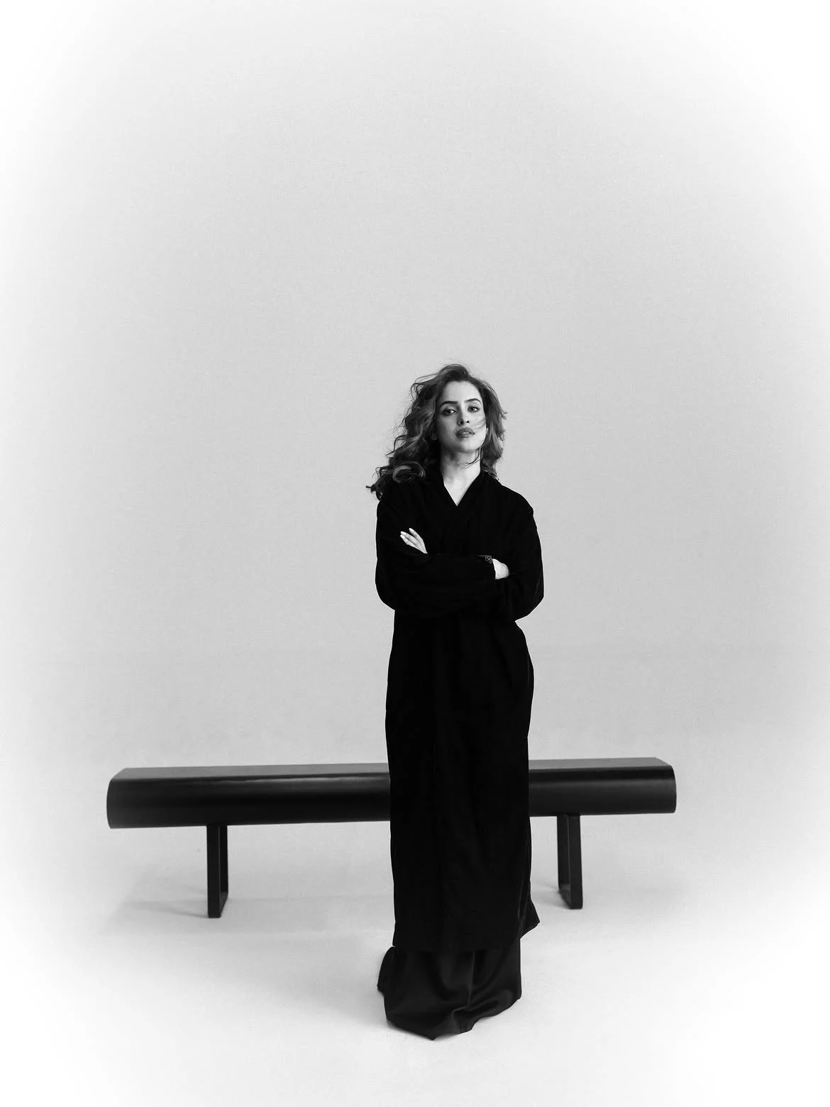 Sanya Malhotra standing in a black and white portrait, wearing a long black coat with arms crossed, looking directly at the viewer. A black minimalist bench is behind her.