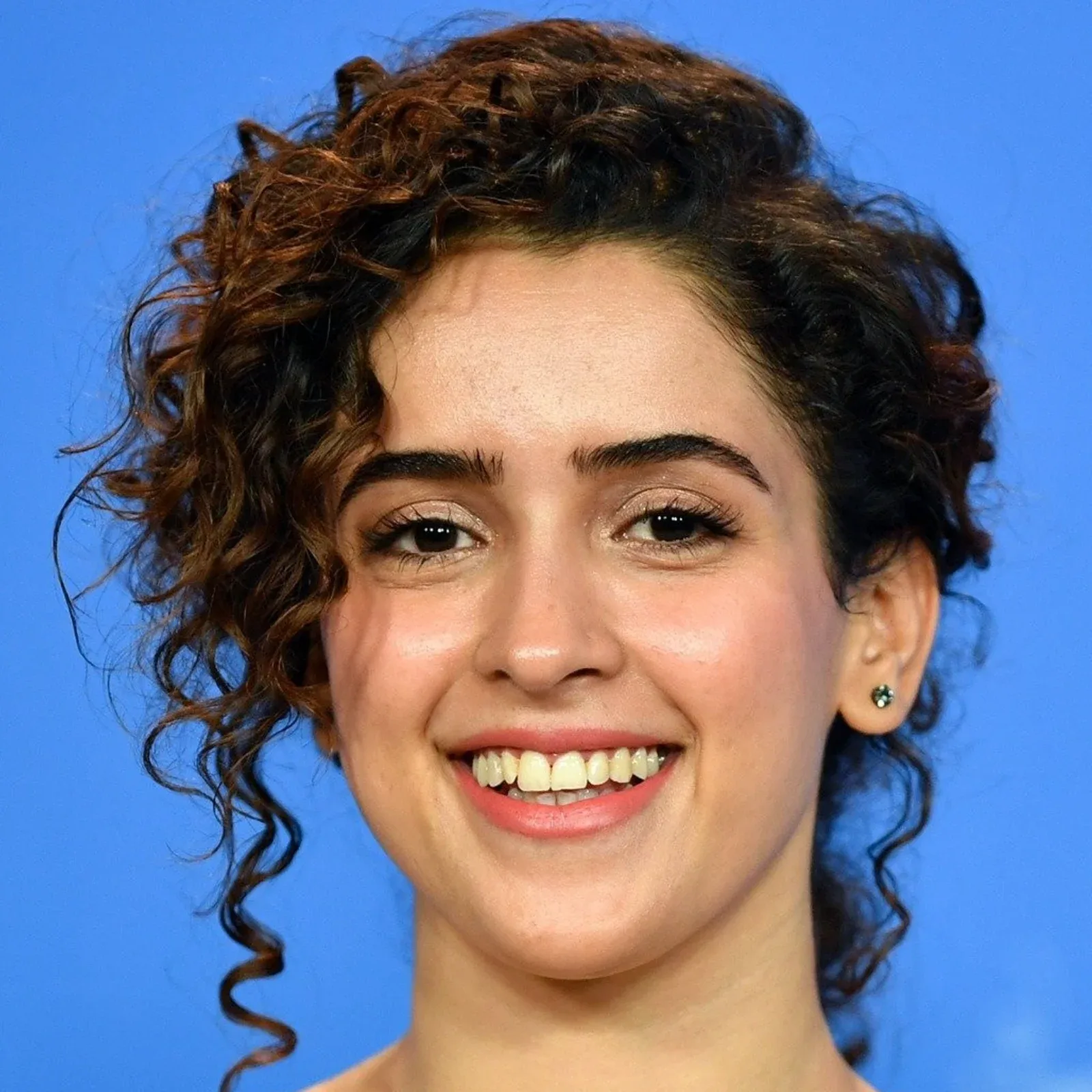 Close-up portrait of Sanya Malhotra smiling, with curly brown hair and a blue background.