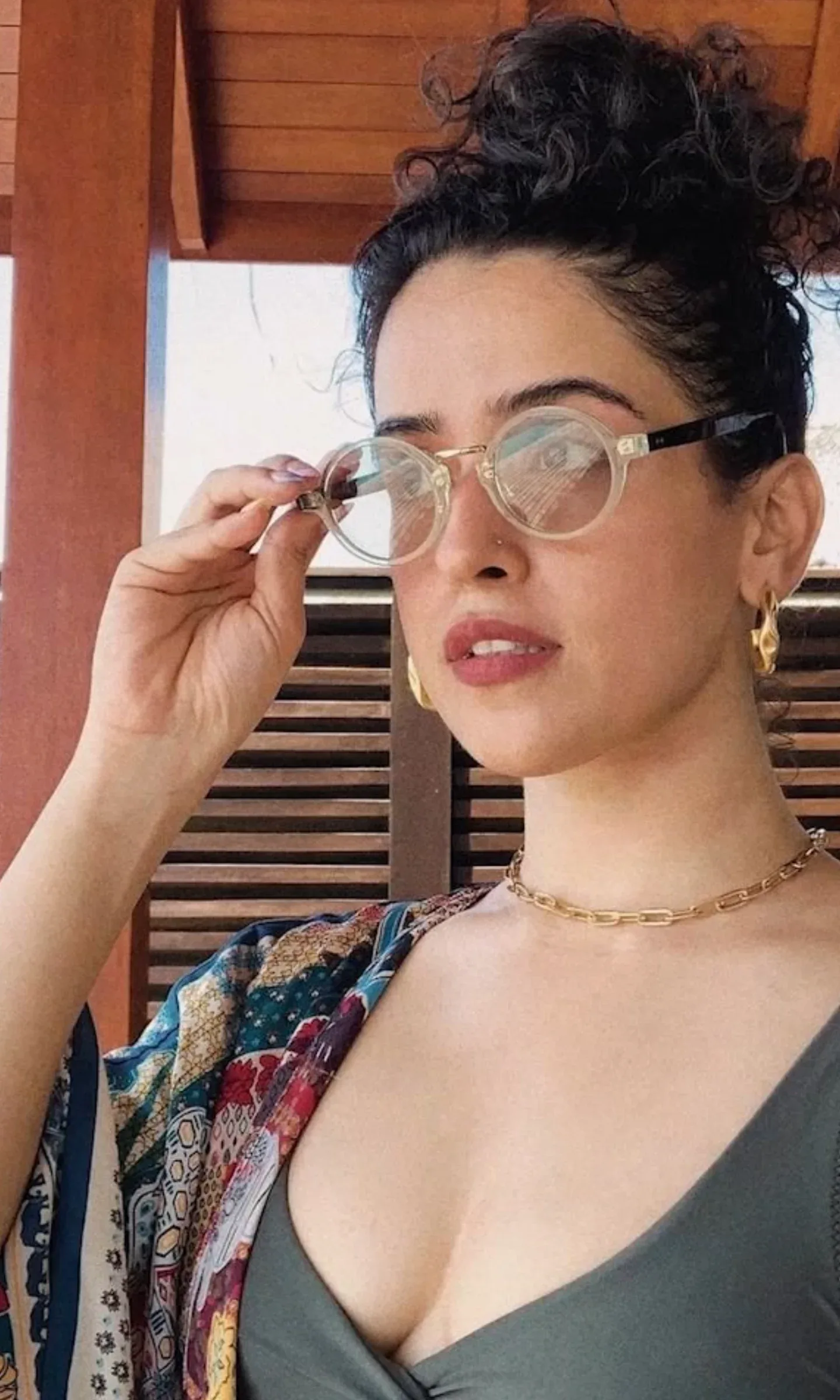 Sanya Malhotra in a close-up, wearing clear-framed round glasses, a dark green top, and a colorful patterned cover-up. Her dark curly hair is in a messy bun, and she has gold jewelry and a nose piercing. Her cleavage is visible.