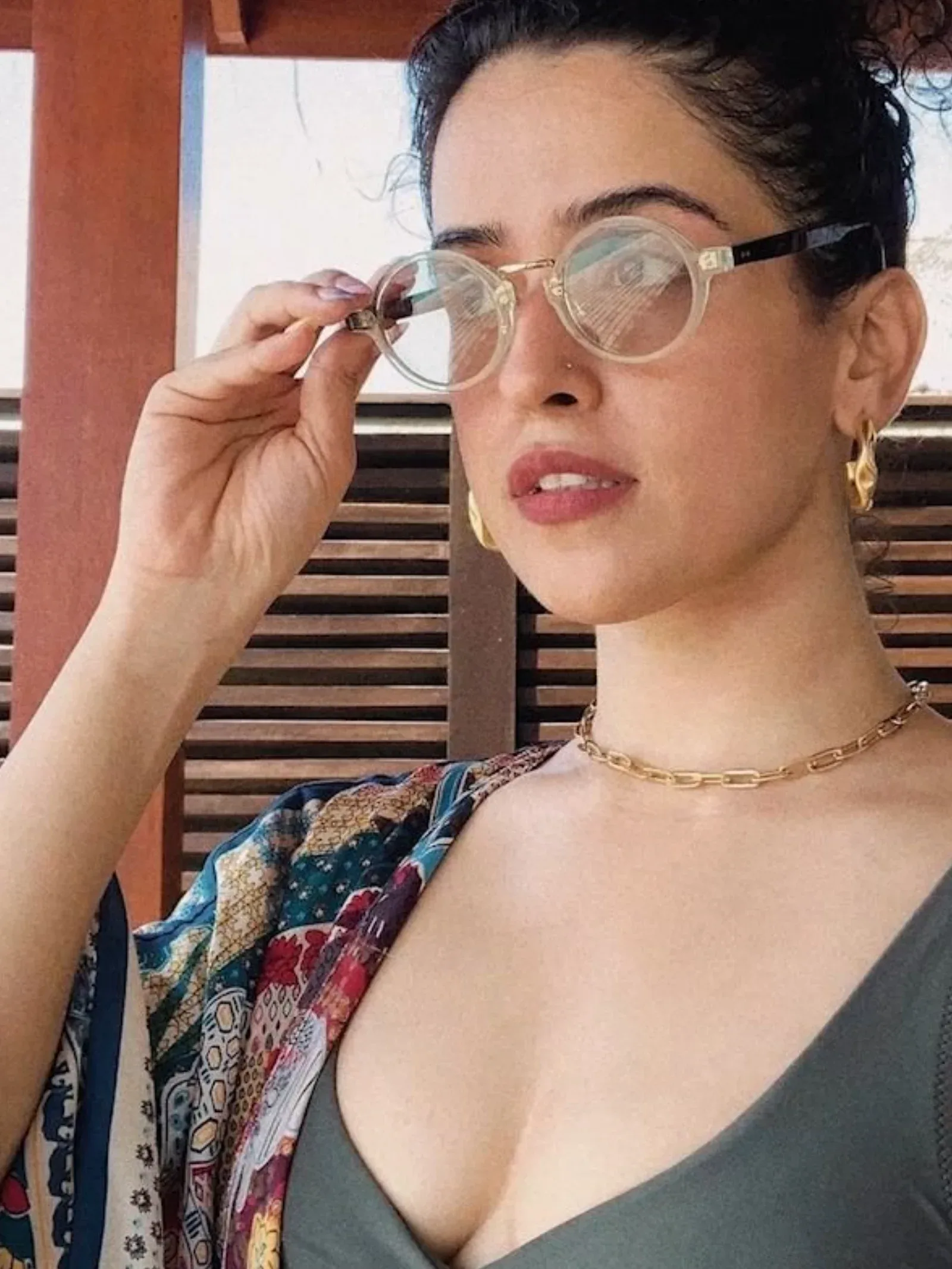 Sanya Malhotra in a close-up, wearing clear round glasses, a dark green top, and a colorful patterned kimono, with a gold chain necklace and earrings.