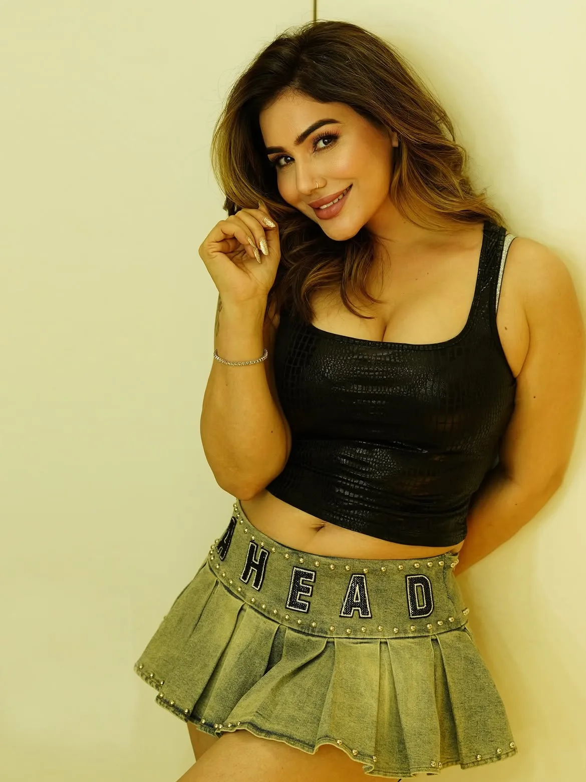 Kangna Sharma smiling, wearing a black textured crop top and a studded pleated denim mini skirt, leaning against a wall.