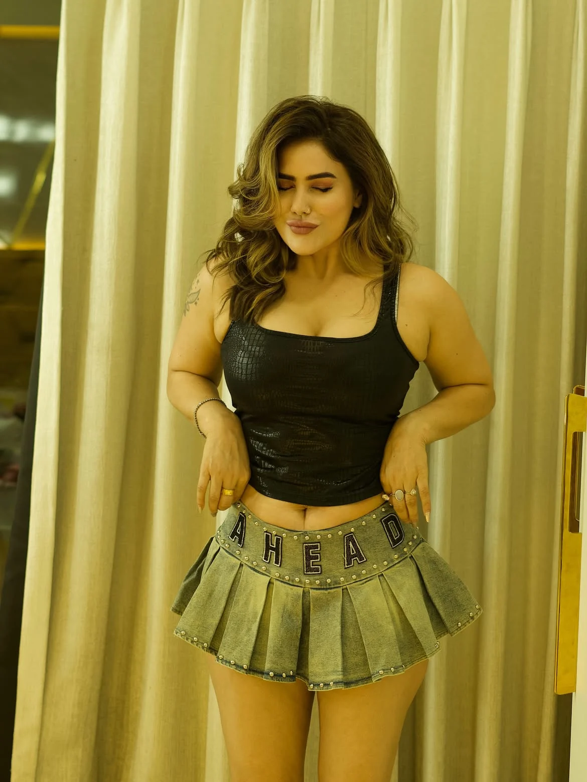 Kangna Sharma in a black textured tank top and a light wash denim pleated mini skirt with 'AHEAD' lettering, hands near her midriff, eyes closed, against a beige curtain.