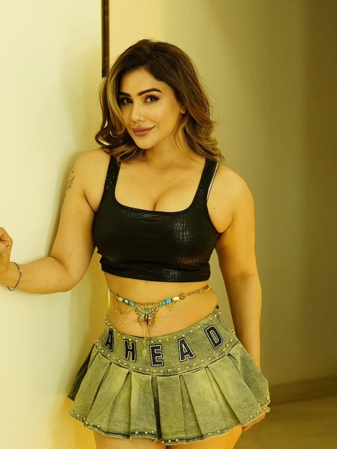 Kangna Sharma in a black textured crop top and light-wash denim pleated mini skirt with a gold waist chain, leaning against a wall and smiling at the camera. Her cleavage, midriff, thighs, and armpits are visible.