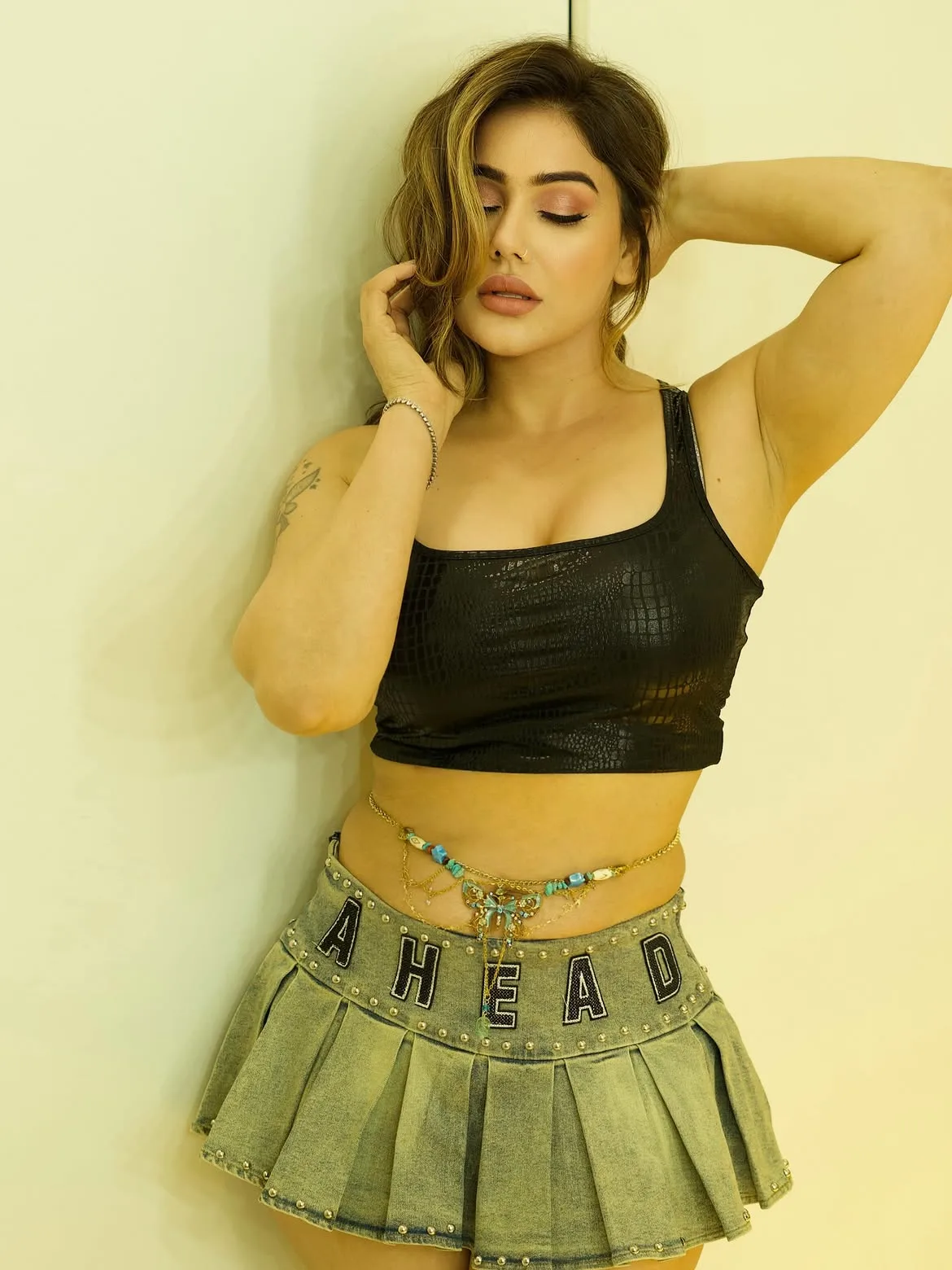 Kangna Sharma posing against a light wall, wearing a black textured crop top, denim pleated mini skirt with 'AHEAD' text, and a beaded waist chain. Her eyes are closed, and her right arm is raised.