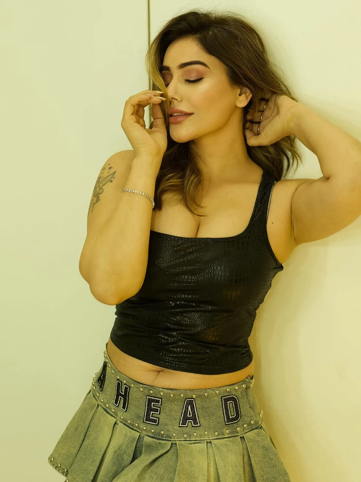 Kangna Sharma posing against a wall, wearing a black textured tank top and a studded denim mini-skirt, with her eyes closed and a hand near her face. Her midriff, cleavage, and armpits are visible.