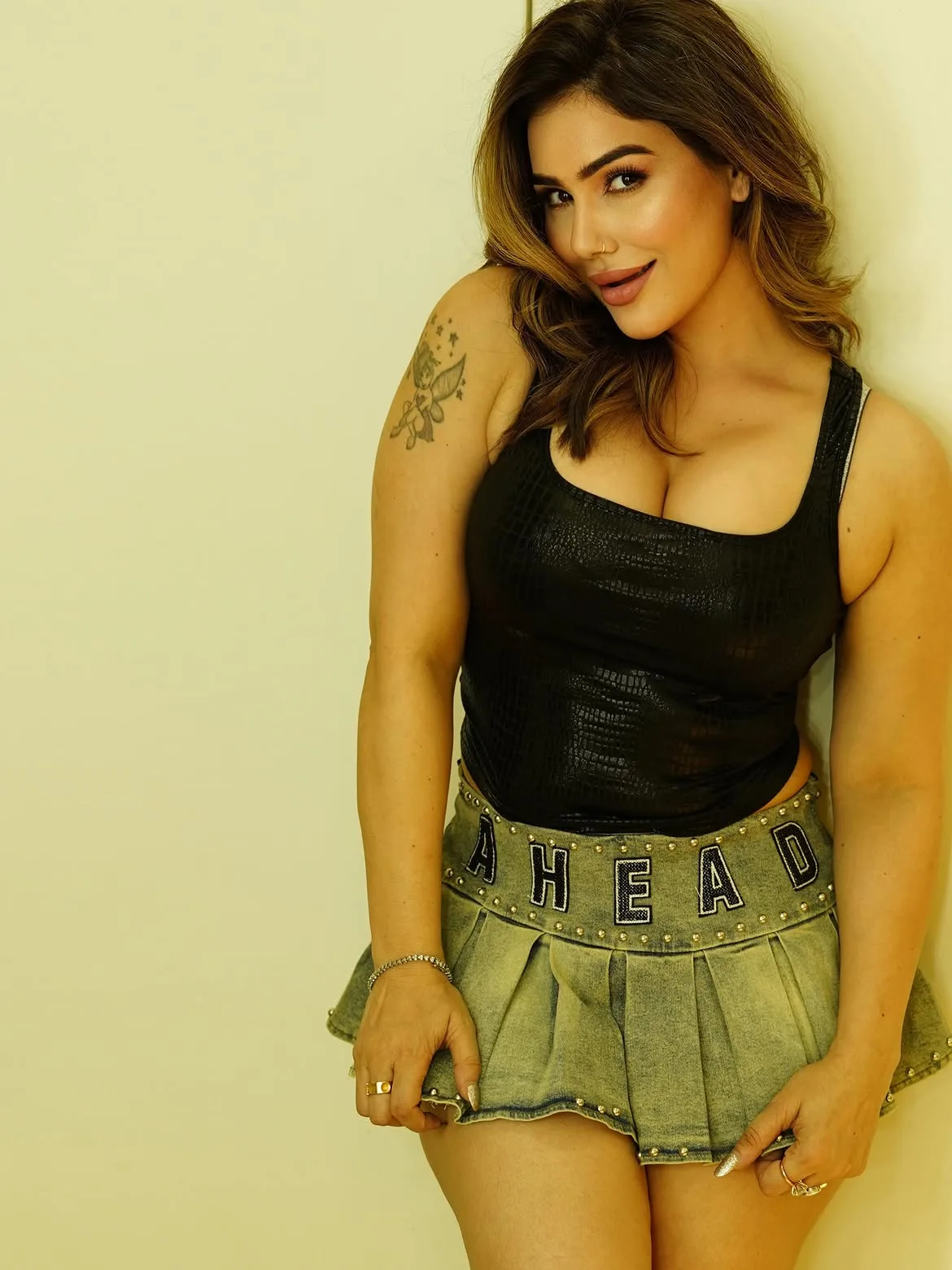 Kangna Sharma smiling, wearing a black textured tank top and a light green pleated denim mini skirt with "AHEAD" on the studded waistband, leaning against a light wall. Her arm tattoo, nose ring, cleavage, midriff, thighs, and armpits are visible.