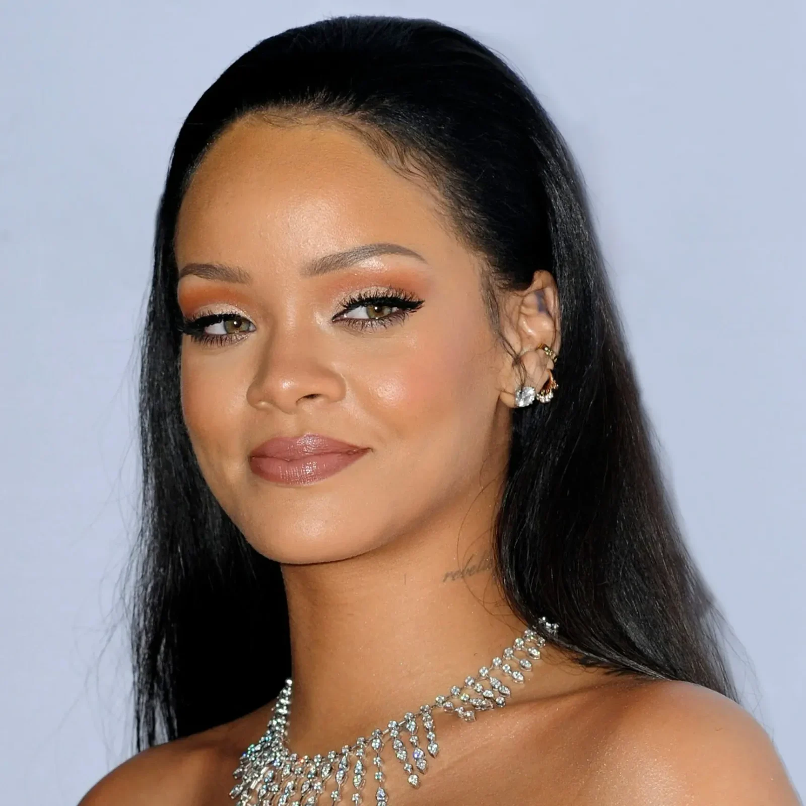 Close-up portrait of Rihanna wearing a strapless top and a large, multi-strand diamond necklace, with her dark hair pulled back and a soft, confident smile.