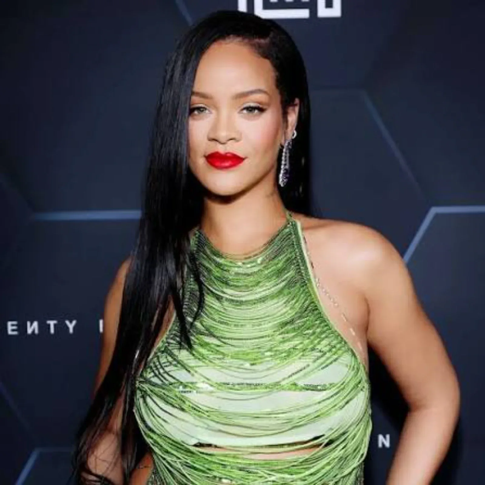 Rihanna in a vibrant green fringed halter top with long black hair and bold red lipstick, posing confidently against a dark background, with a hint of midriff visible.