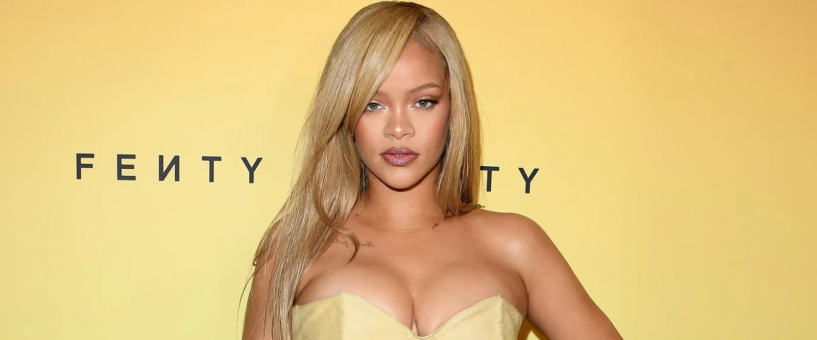 Rihanna with long blonde hair and bangs, wearing a strapless light yellow dress, looking directly at the camera with a confident expression. The FENTY logo is visible in the yellow background.