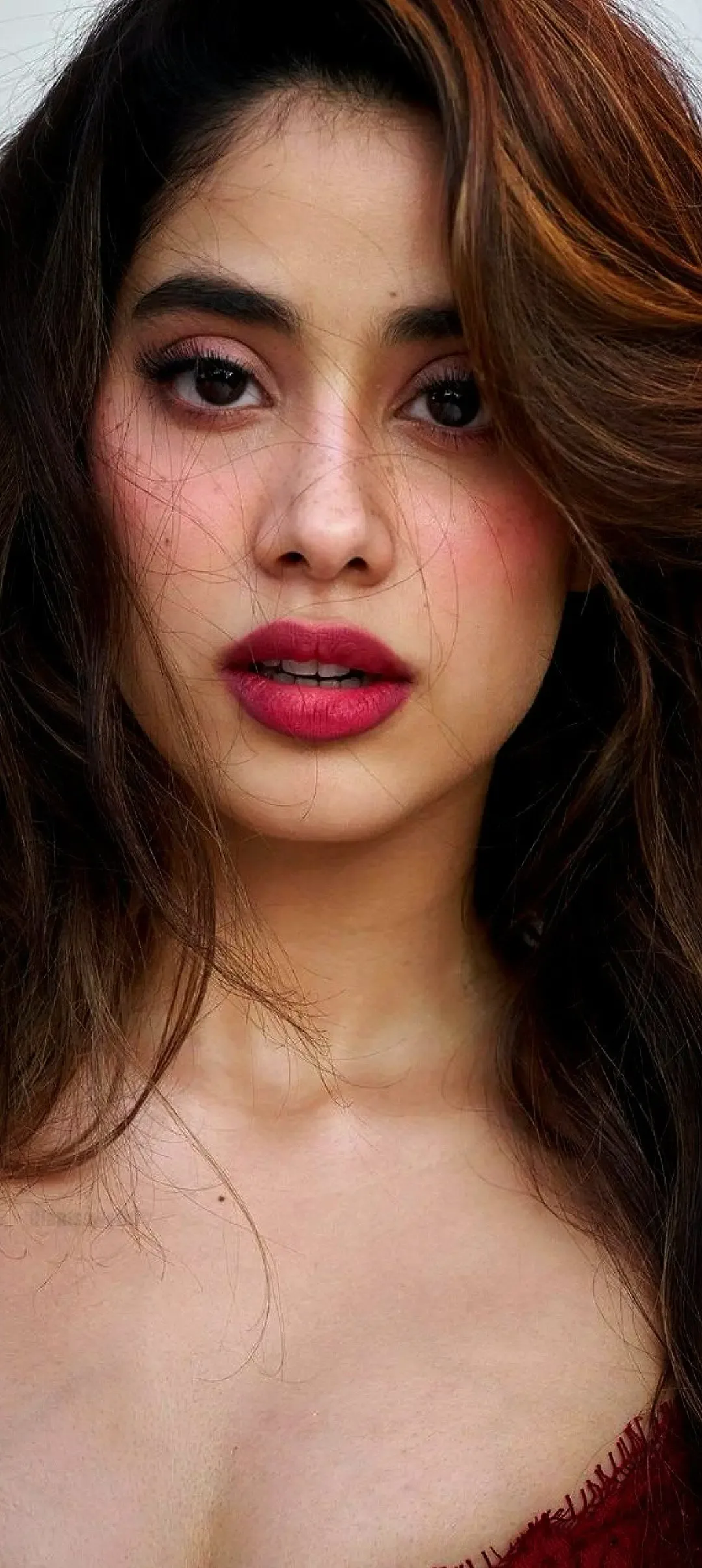 Close-up portrait of Janhvi Kapoor with long brown hair, bold red lipstick, and expressive eyes, wearing a red top.