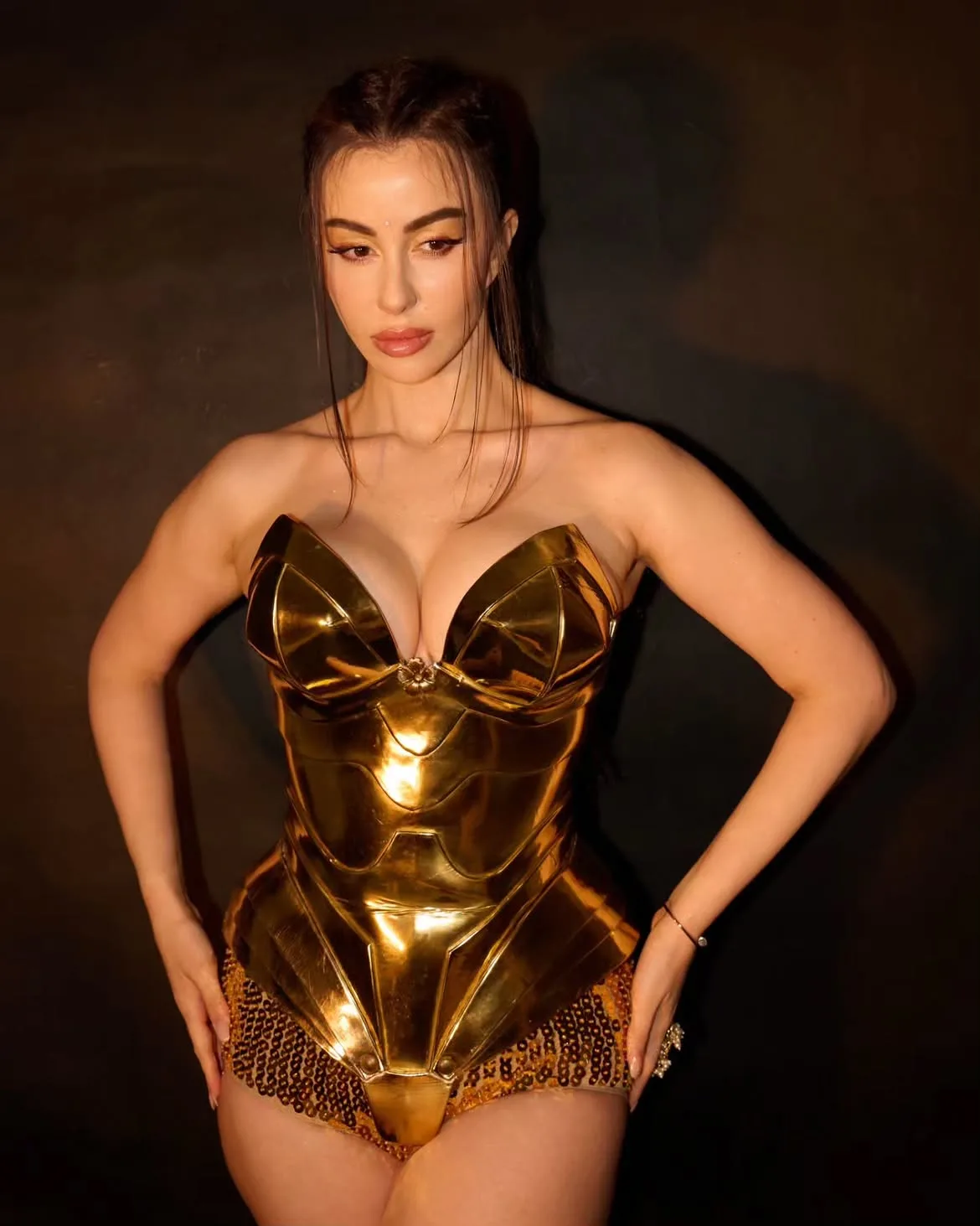 Giorgia Andriani in a golden, strapless, armored-style bodysuit with sequined bottoms, posing with hands on hips against a dark background, showcasing cleavage and thighs.
