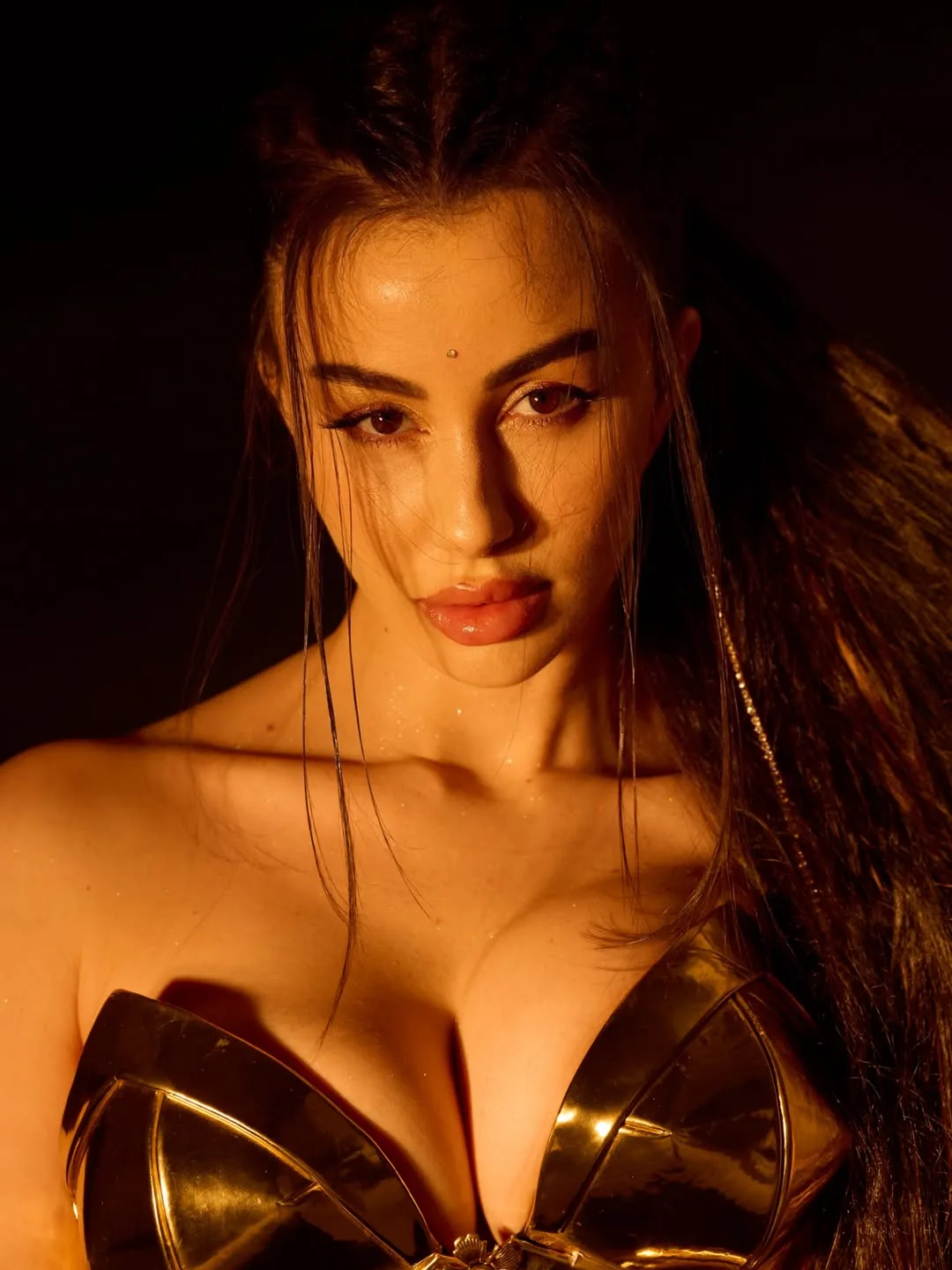 Giorgia Andriani in a golden strapless top, looking intensely at the camera with long dark hair and prominent cleavage, bathed in warm light.