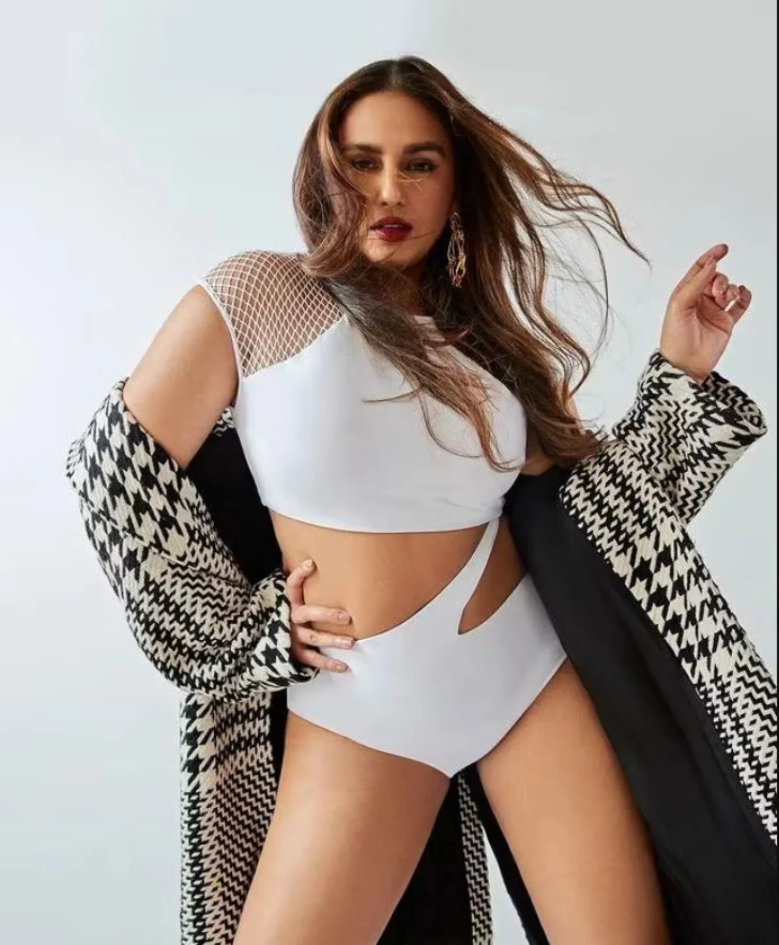 Huma Qureshi posing confidently in a white cutout swimsuit and houndstooth coat showcasing her midriff and thighs.