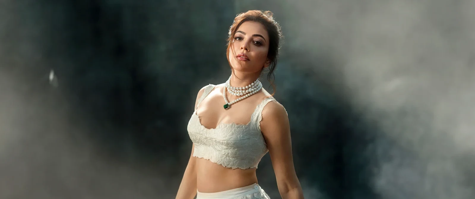 Kajal Aggarwal wearing a white embroidered crop top and multi-layered pearl necklace in a smoky studio photo.
