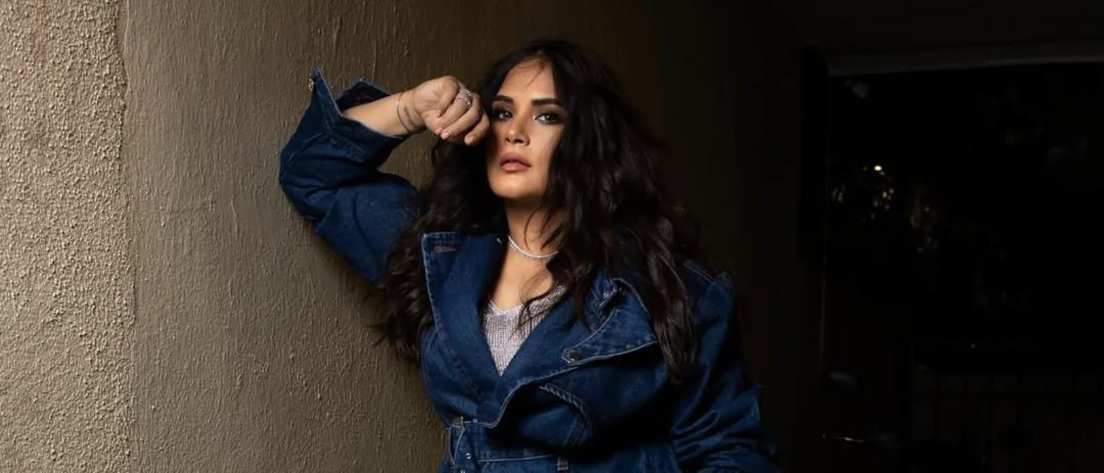 Richa Chadha leaning against a wall wearing a denim jacket and silver top