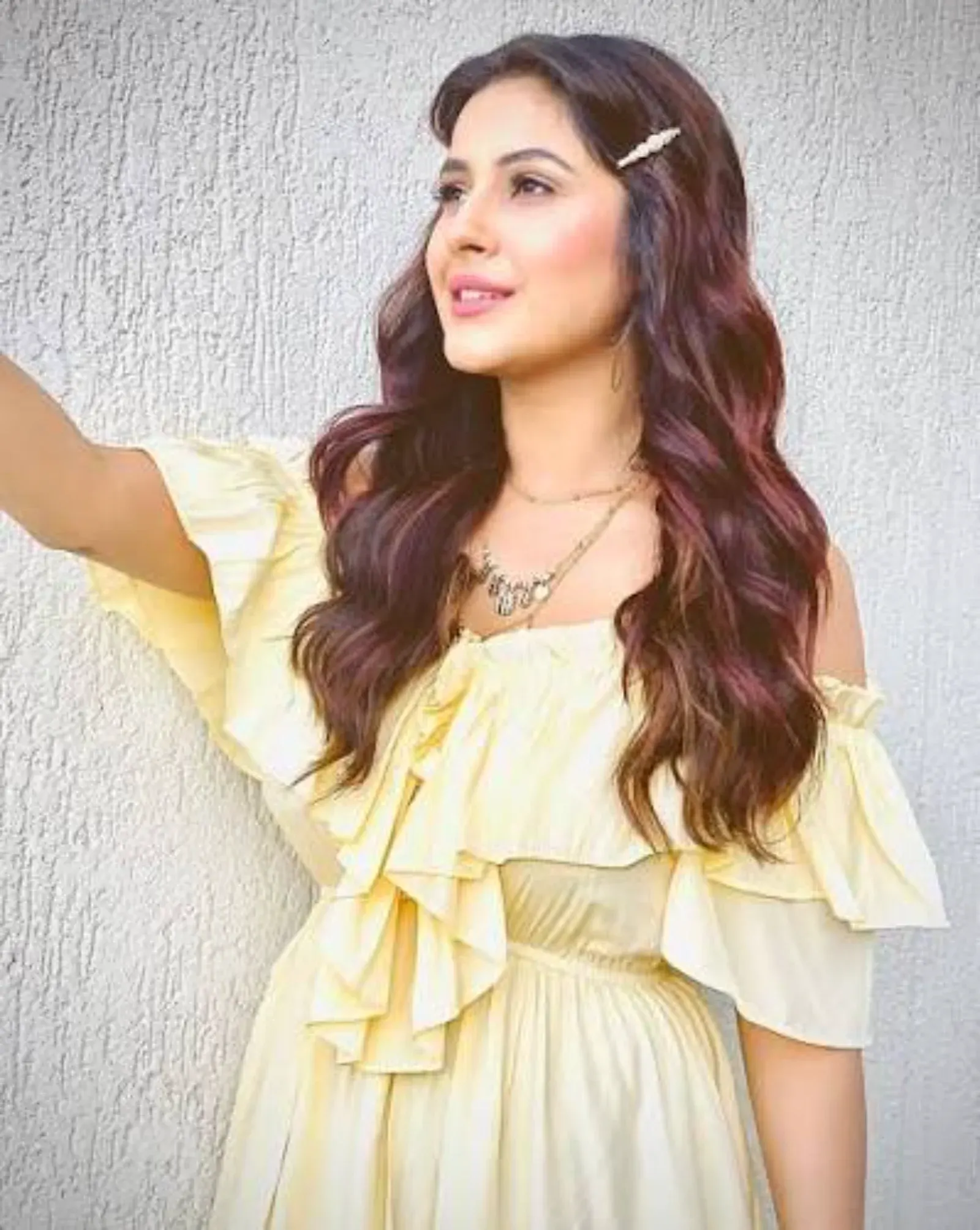 Shehnaaz Gill posing in a yellow off-the-shoulder ruffled dress with wavy hair