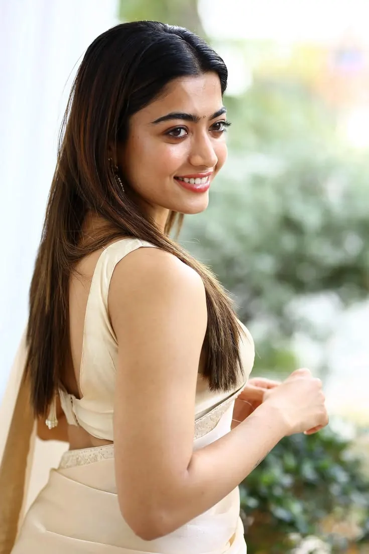 Rashmika Mandanna posing gracefully in an elegant cream saree and sleeveless blouse with a warm smile