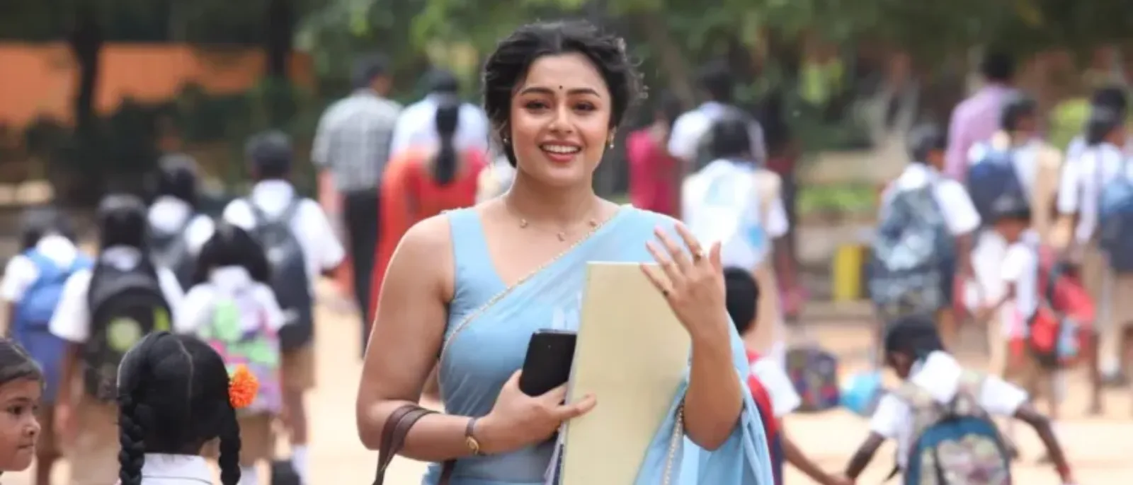 Mokksha as Meenu Miss walking through a school campus wearing a light blue sleeveless saree in Gatta Kusthi 2