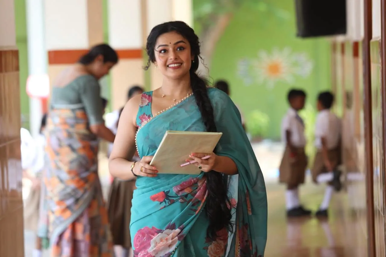 Mokksha smiling in a teal floral saree holding a folder in a school corridor