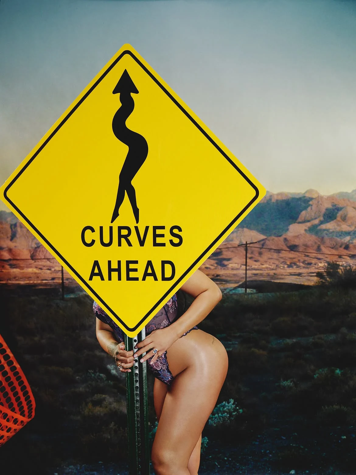 Rihanna posing behind a yellow Curves Ahead road sign wearing floral lace lingerie