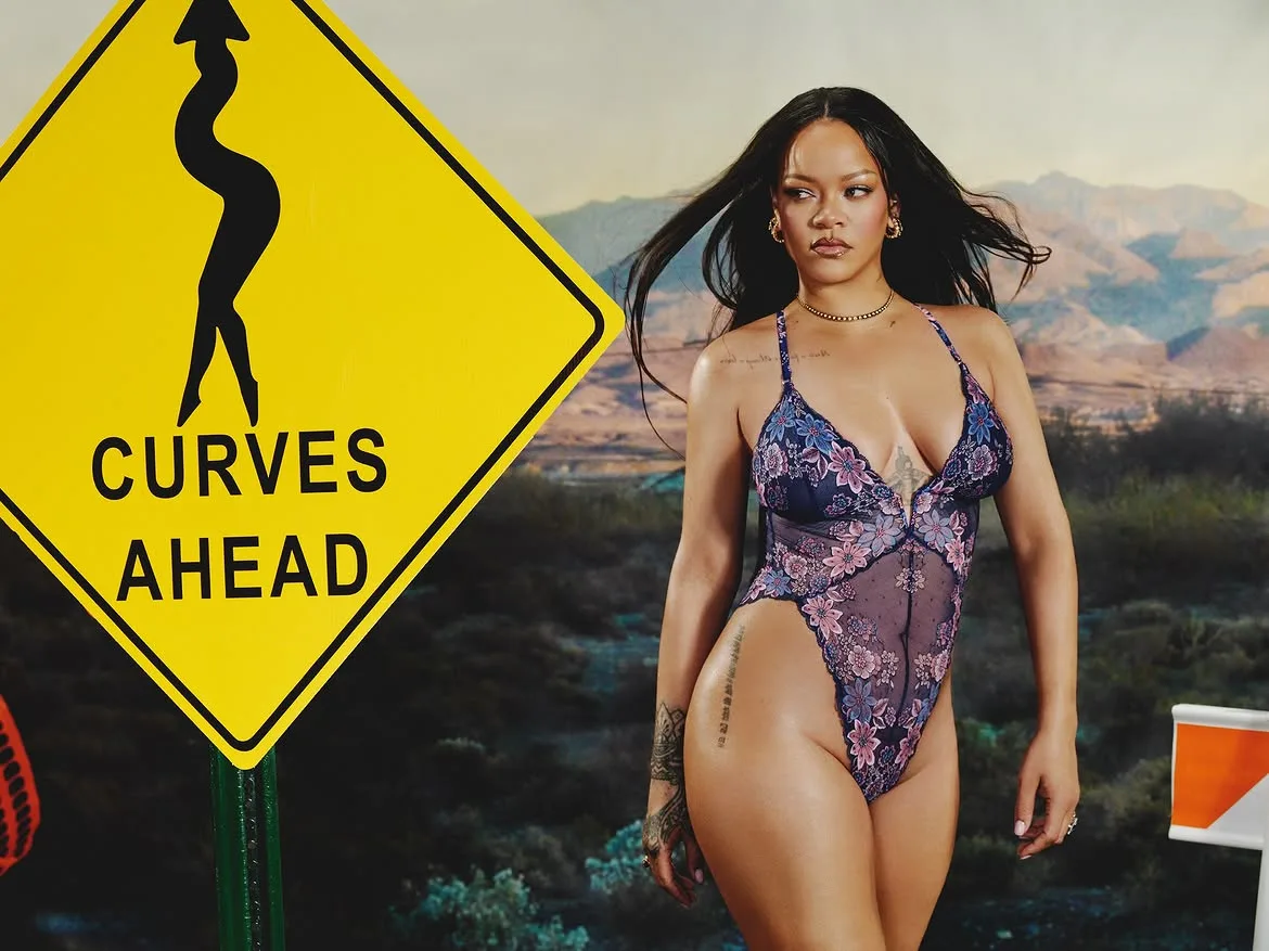 Rihanna wearing a sheer floral lace bodysuit posing beside a yellow Curves Ahead road sign in a desert