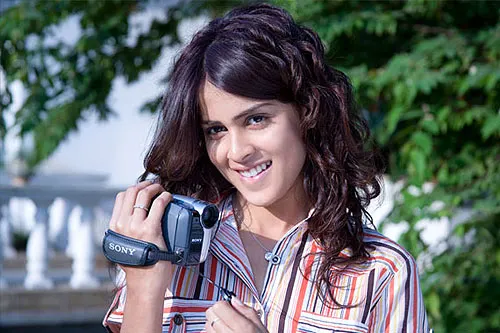 Genelia D'Souza smiling while holding a video camera in a movie still from Jaane Tu Ya Jaane Na