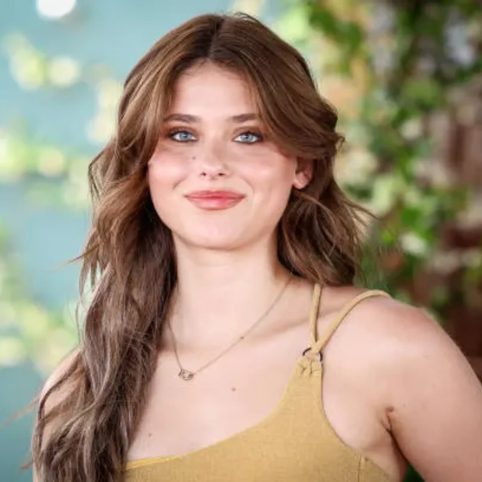 Nicole Wallace posing with long wavy hair and blue eyes wearing a yellow strap top and gold necklace